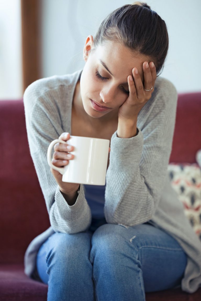 Depressed young woman having headache while drinking coffee on sofa at home.